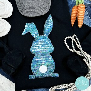 May include: Black sweatshirt with a sequined blue bunny design. The bunny has a white fluffy tail. The image also includes a grey hat, white shoes, a string of pearls, and decorative carrots.