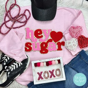 May include: A light pink sweatshirt with the words "hey sugar" in red and pink letters with gold trim. The image also includes a black baseball cap, a pair of black sneakers, and a decorative sign that says "XOXO".