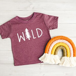 Może przedstawiać: Bordowy t-shirt z kr&oacute;tkim rękawem z napisem "WILD" w białych literach i białą sylwetką sosny. W tle znajduje się tęczowa makrama.