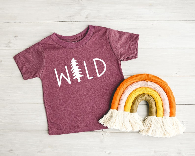 Może przedstawiać: Bordowy t-shirt z kr&oacute;tkim rękawem z napisem "WILD" w białych literach i białą sylwetką sosny. W tle znajduje się tęczowa makrama.