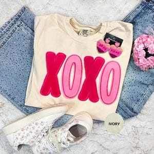 Op de afbeelding: Een ivoorkleurig T-shirt met de letters XOXO in roze en een donkerroze omtrek. De afbeelding bevat lichtblauwe jeans, bloemen sneakers en hartvormige oorbellen. Een klein roze bloemenaccessoire is ook inbegrepen.
