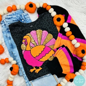 May include: A black and pink turkey graphic on a black shirt with a leopard print pattern. The shirt is layered over a black and orange striped sweater with pink accents. The sweater is decorated with a garland of white and orange pom-poms.