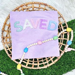 May include: A lavender t-shirt with the word "SAVED" in pastel-colored, textured letters. The shirt is displayed on a woven basket, with a beaded garland featuring colorful egg-shaped beads.