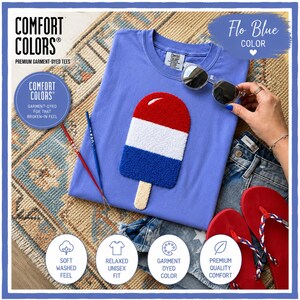 May include: A light blue Comfort Colors t-shirt with a red, white, and blue popsicle design. The shirt is part of a collection of premium garment-dyed tees. The image also includes sunglasses, denim shorts, and red flip-flops, suggesting a summer theme.
