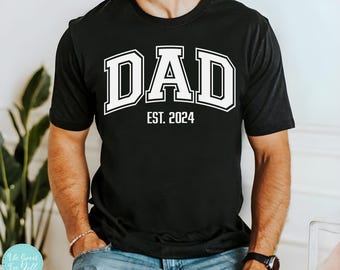 Puede incluir: Una camiseta negra con texto blanco que dice "DAD EST. 2024". El texto está en una fuente grande y en negrita y está diseñado para parecerse al logotipo de un equipo deportivo universitario.