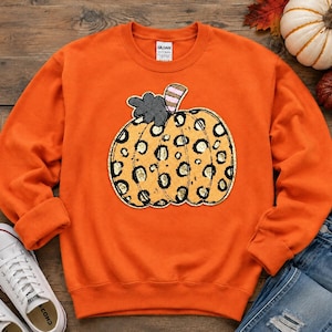 Puede incluir: Una sudadera naranja de manga larga y cuello redondo con un gran gráfico central de una calabaza con estampado de leopardo dorado y negro, un detalle de hoja gris oscuro y un pequeño tallo a rayas. La etiqueta Gildan es visible en el cuello.