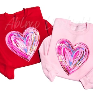 May include: Two sweatshirts, one red and one pink, each featuring a large heart design. The heart is filled with a colorful, textured pattern of pink, blue, and gold. The text "Ablnco L" is visible on the red sweatshirt.