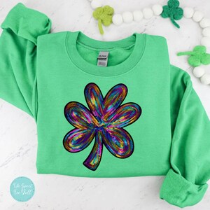 Puede incluir: Una sudadera verde con un gran diseño de trébol con lentejuelas en colores arcoíris. El trébol es el punto focal, con un contorno detallado. La sudadera tiene mangas largas y cuello redondo.