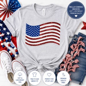 May include: A light gray t-shirt featuring a sequined American flag design. The shirt has a crew neck and short sleeves. The image includes white sneakers, denim shorts, and patriotic decorations. The shirt is labeled as soft, lightweight, and unisex.
