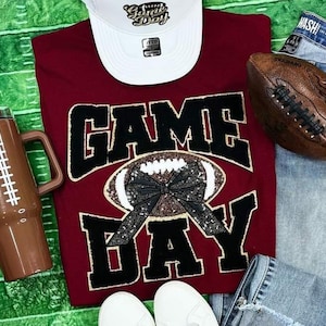 Peut inclure: Un t-shirt bordeaux avec les mots "GAME DAY" en noir, avec un motif de ballon de football avec un nœud noir. Une casquette de baseball blanche avec le texte "Game Day", un ballon de football marron, un gobelet marron et un jean complètent l'ensemble.
