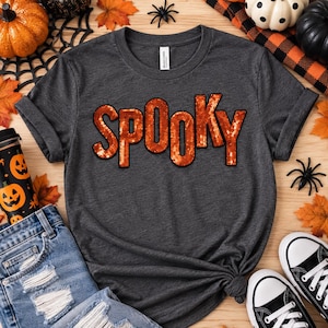 May include: A dark heather gray short-sleeve t-shirt featuring the word SPOOKY printed in large, orange, sequin-style letters across the chest. The shirt has a crew neckline and a standard fit, with a white brand label visible at the inner collar.