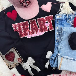 May include: A black sweatshirt with the text "HEART breaker" in red and pink sequins, alongside light wash denim shorts, a pink corduroy cap, and heart decorations. The image has a Valentine's Day theme.