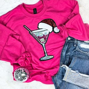 Puede incluir: Una sudadera rosa brillante con un diseño de copa de cóctel con lentejuelas y un gorro de Papá Noel. El gráfico incluye cubitos de hielo y una pajita. La sudadera se combina con vaqueros azules y una bola de discoteca.