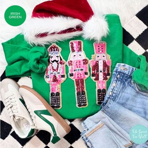 May include: A green sweatshirt with three pink and red nutcracker appliques, paired with light-wash denim jeans, white and green sneakers, and a Santa hat. The sweatshirt has the text "Irish Green" in a green circle.