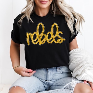 May include: A black short-sleeved t-shirt featuring the word rebels printed in a gold glitter script font across the chest. The text has a distinct outline and a textured, sparkling appearance that contrasts against the solid black fabric.