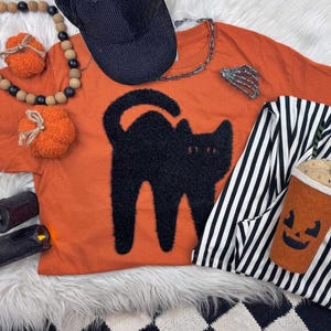 May include: Orange t-shirt with a black cat design, a black corduroy baseball cap, and a black and white striped jacket. Decorative orange pumpkin ornaments and a Halloween-themed coffee cup are also present.