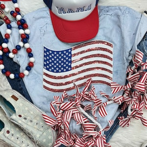 Puede incluir: Una camiseta azul claro con un diseño de bandera americana de lentejuelas rojas, blancas y azules. La camiseta también tiene una gorra de béisbol roja con la palabra "MAMA" en letras blancas. La camiseta está rodeada de cintas rojas y blancas.
