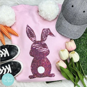 May include: A light pink sweatshirt with a sequined bunny design. The image also includes a gray baseball cap, black sneakers, white pom-poms, carrot decorations, and tulips. The sweatshirt is a festive Easter-themed item.