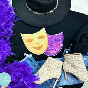May include: Black sweatshirt featuring gold and purple theatre masks. The image includes a black hat with silver trim, denim jeans, and beige patterned ankle boots. A purple feather boa is draped on the side.