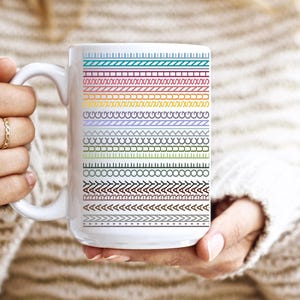 May include: White ceramic mug featuring a series of colorful, patterned borders. The designs include lines, zigzags, and geometric shapes in various colors. The mug is held by a person wearing a cream-colored sweater.