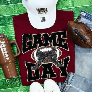 May include: A maroon t-shirt with the text "Game Day" in black and gold lettering. A football with a black and gold bow is centered on the shirt. A white baseball cap with the text "Game Day" is on top of the shirt. A brown football and a brown insulated tumbler are also in the image.