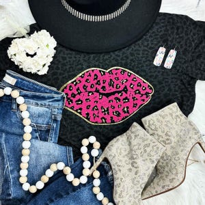 May include: A fashion flat lay featuring a black hat with a rhinestone band, a black top with a pink leopard print lip design, blue jeans, and leopard print high-heeled ankle boots. A white beaded garland and earrings are also included.