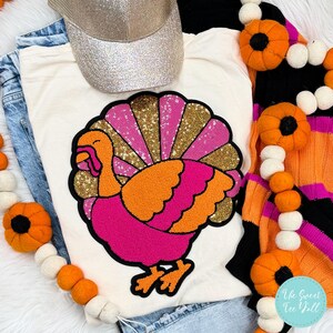 May include: A white t-shirt featuring a pink and orange sequined turkey applique. The turkey has a gold glitter tail and a pink and orange body. The shirt is surrounded by a garland of orange and white pumpkins.