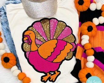 Op de afbeelding: Een wit T-shirt met een roze en oranje pailletten-kalkoen-applicatie. De kalkoen heeft een gouden glitterstaart en een roze en oranje lichaam. Het shirt is omringd door een krans van oranje en witte pompoenen.
