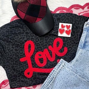 Op de afbeelding: Een zwart t-shirt met luipaardprint met het woord "Love" in rode letters, gecombineerd met een lichtblauwe jeans en een rood-zwarte geruite baseballcap. Een paar rode hartvormige oorbellen maakt de outfit compleet.