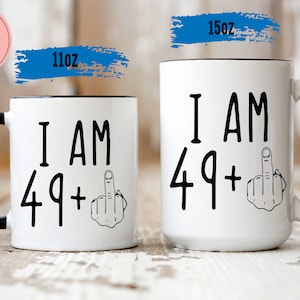 May include: Two white ceramic coffee mugs with black handles. The mugs have black text that reads "I AM 49+" with a middle finger emoji. The smaller mug is 11oz and the larger mug is 15oz.