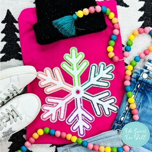 Pode incluir: Uma camisa rosa choque de manga comprida com um grande desenho de floco de neve colorido. A imagem também inclui um gorro preto com uma borla turquesa, tênis brancos, jeans azuis e uma guirlanda de contas coloridas. O texto "The Sweet Tee Yell" é visível.