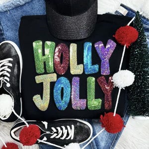 Può includere: Felpa nera con le parole "HOLLY JOLLY" in lettere paillettate colorate. L'immagine include scarpe da ginnastica nere, un berretto da baseball nero glitterato e decorazioni natalizie come un piccolo albero di Natale e pompon.