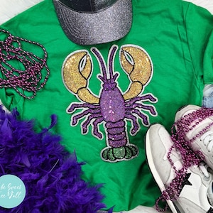 May include: A green t-shirt with a sequined purple lobster design and gold claws. The image includes a silver glitter baseball cap, purple beaded necklaces, a purple feather boa, and white trainers.