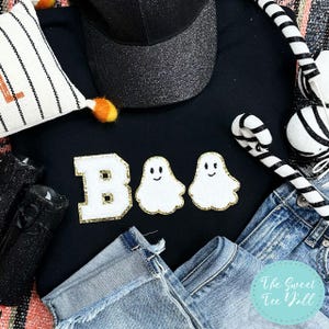 May include: A black sweatshirt featuring the word "BOO" in white and gold, with two ghost appliques. A black glitter cap, denim jeans, and Halloween decorations are arranged around the sweatshirt.