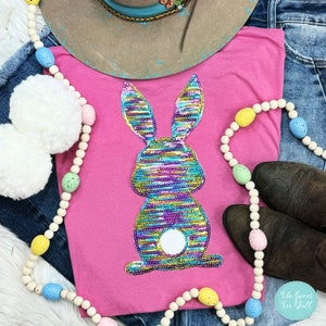 May include: A pink t-shirt with a sequined bunny design in blue, green, purple, and gold. The shirt is styled with denim jeans, a cowboy hat, and Easter egg garland. The Sweet Tee Yall logo is visible.