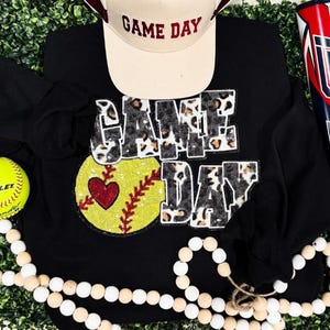 Może przedstawiać: Czarna koszulka z długim rękawem z cekinową grafiką do softballu i napisem "GAME DAY". Na górze beżowa czapka z daszkiem z napisem "GAME DAY". Widoczna jest również żółta piłka do softballu i ozdobne koraliki.