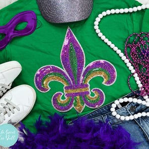 May include: A green t-shirt featuring a sequined fleur-de-lis design in purple, green, and gold. The image includes a purple mask, a silver glitter hat, white trainers, and beaded necklaces. A purple feather boa and denim jeans are also present.