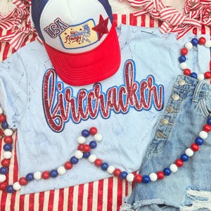 May include: A patriotic ensemble featuring a light blue t-shirt with the word "Firecracker" in red and blue glitter, a red, white, and blue trucker hat, and denim shorts. A red star and USA patch adorn the hat. A beaded garland in red, white, and blue completes the look.