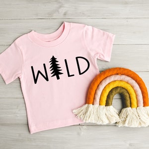 Może przedstawiać: R&oacute;żowy t-shirt z nadrukiem słowa "WILD" w kolorze czarnym z grafiką sosny. Ręcznie robiona wisząca ozdoba w kształcie tęczy z białymi frędzlami.