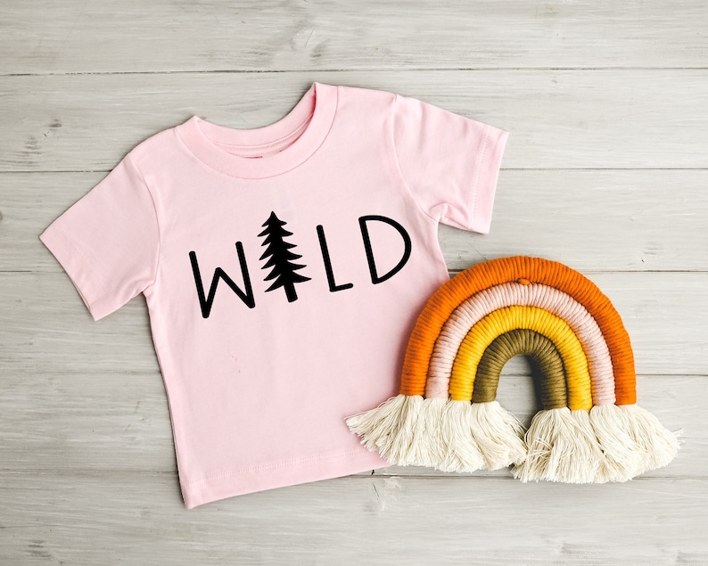 Może przedstawiać: R&oacute;żowy t-shirt z nadrukiem słowa "WILD" w kolorze czarnym z grafiką sosny. Ręcznie robiona wisząca ozdoba w kształcie tęczy z białymi frędzlami.