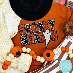 Może przedstawiać: Pomarańczowa koszulka z napisem "Spooky Season" w cekinowym wzorze, białym duchem i dyniowymi akcentami. Obraz zawiera czarny kapelusz, kremowy kardigan oraz girlandę z filcowych dyń i białych kulek.
