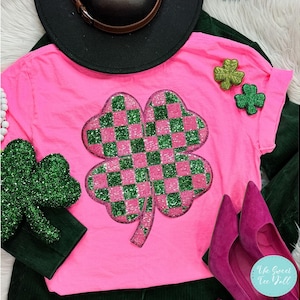 May include: Pink t-shirt with a large green and pink four-leaf clover with a checkered pattern. The clover is made of sequins and is a sparkly embellishment. The shirt has a distressed look and is a casual style.