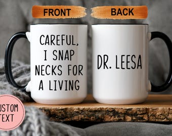 Pode incluir: Caneca de cerâmica branca com alça preta. A frente da caneca diz "Careful, I snap necks for a living". O verso da caneca diz "Dr. Leesa".