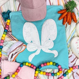 May include: A turquoise t-shirt featuring a white bunny ear design, a pink corduroy cap, and a bunch of carrots. A colourful beaded garland and striped fabric are also visible. The design is perfect for Easter.