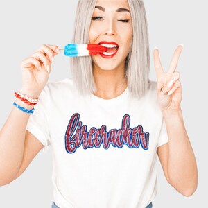 May include: A white t-shirt with the word "firecracker" written in red and blue glitter lettering. The model is wearing the shirt and holding a red, white, and blue popsicle.