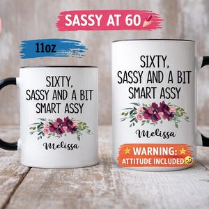 May include: A white ceramic mug with a black handle and the text "But First, Coffee" in a bold, black font. The mug is set against a blurred background, suggesting a cozy setting.