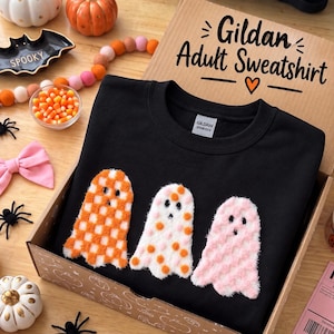 May include: A black crewneck sweatshirt featuring three fuzzy ghost appliques in a row. The left ghost has an orange and white checkered pattern, the middle ghost has orange polka dots, and the right ghost is solid pink. The sweatshirt is in a box.