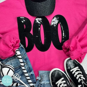 May include: A bright pink sweatshirt with the word "BOO" in large, black sequined letters. The image also includes a black glitter baseball cap, denim shorts, black sequined sneakers, and decorative black and white striped items.