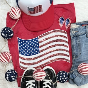 May include: Red, white, and blue patriotic themed t-shirt with a sequined American flag design. The t-shirt is paired with a white baseball cap with a red, white, and blue American flag patch.  The t-shirt is also paired with a pair of black sneakers and a pair of blue jeans.