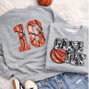Könnte beinhalten: Zwei graue Sweatshirts mit Basketball-Designs. Eines zeigt die Zahl "18" in Orange und Schwarz, die einem Basketball ähnelt. Das andere sagt "GAME DAY" mit einer Basketballgrafik und einem Herz. Ein Basketball und Jeansshorts sind ebenfalls zu sehen.